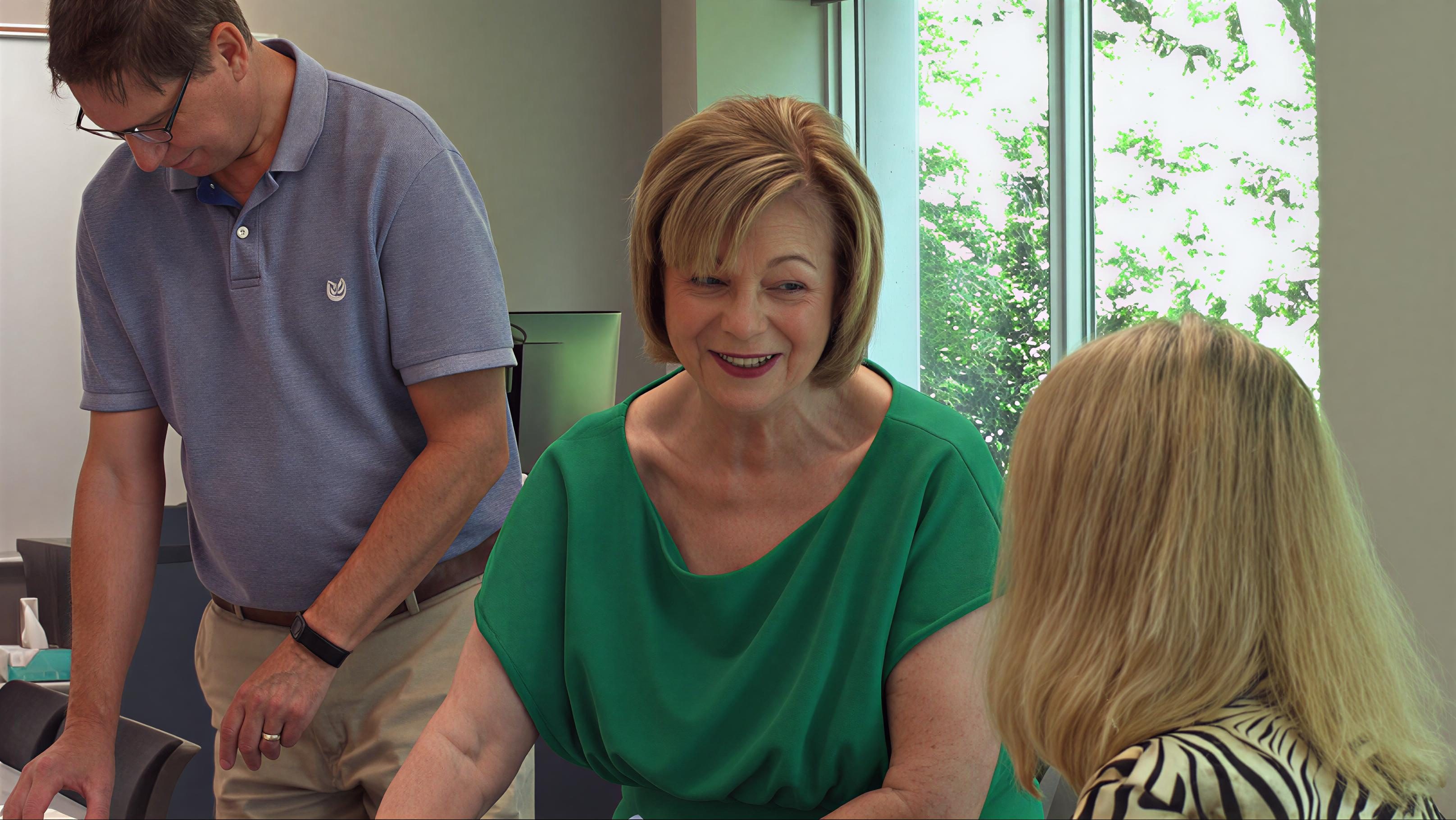 A woman in a green top smiling and interacting with two others indoors.
